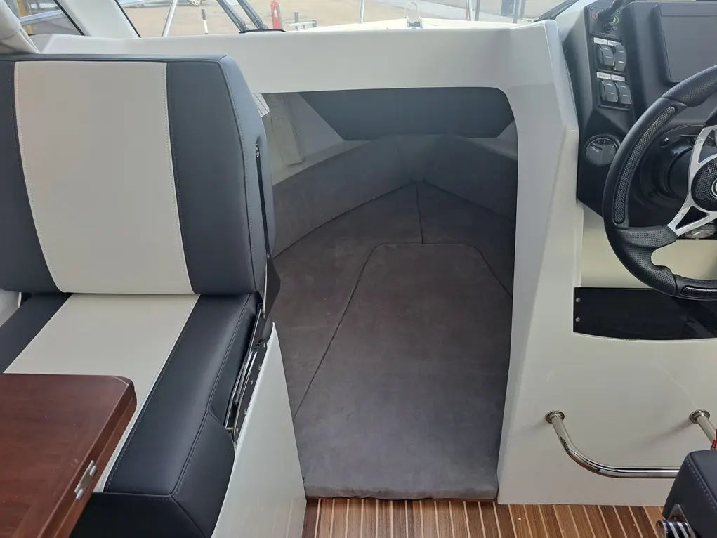 Interior view of a boat featuring a padded seat, steering wheel, and a grey lounging area with wood flooring.
